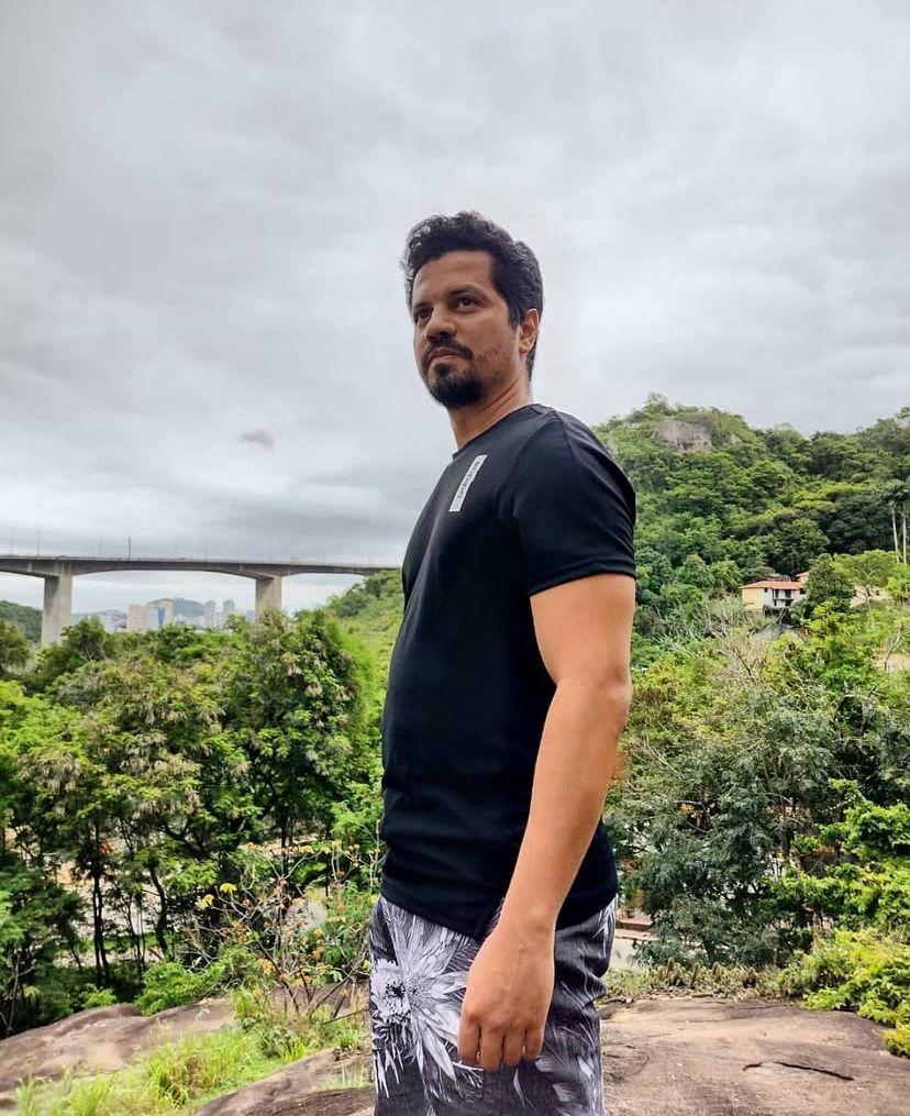 Manoel Ednaldo em ambiente ao ar livre, com paisagem e ponte ao fundo.